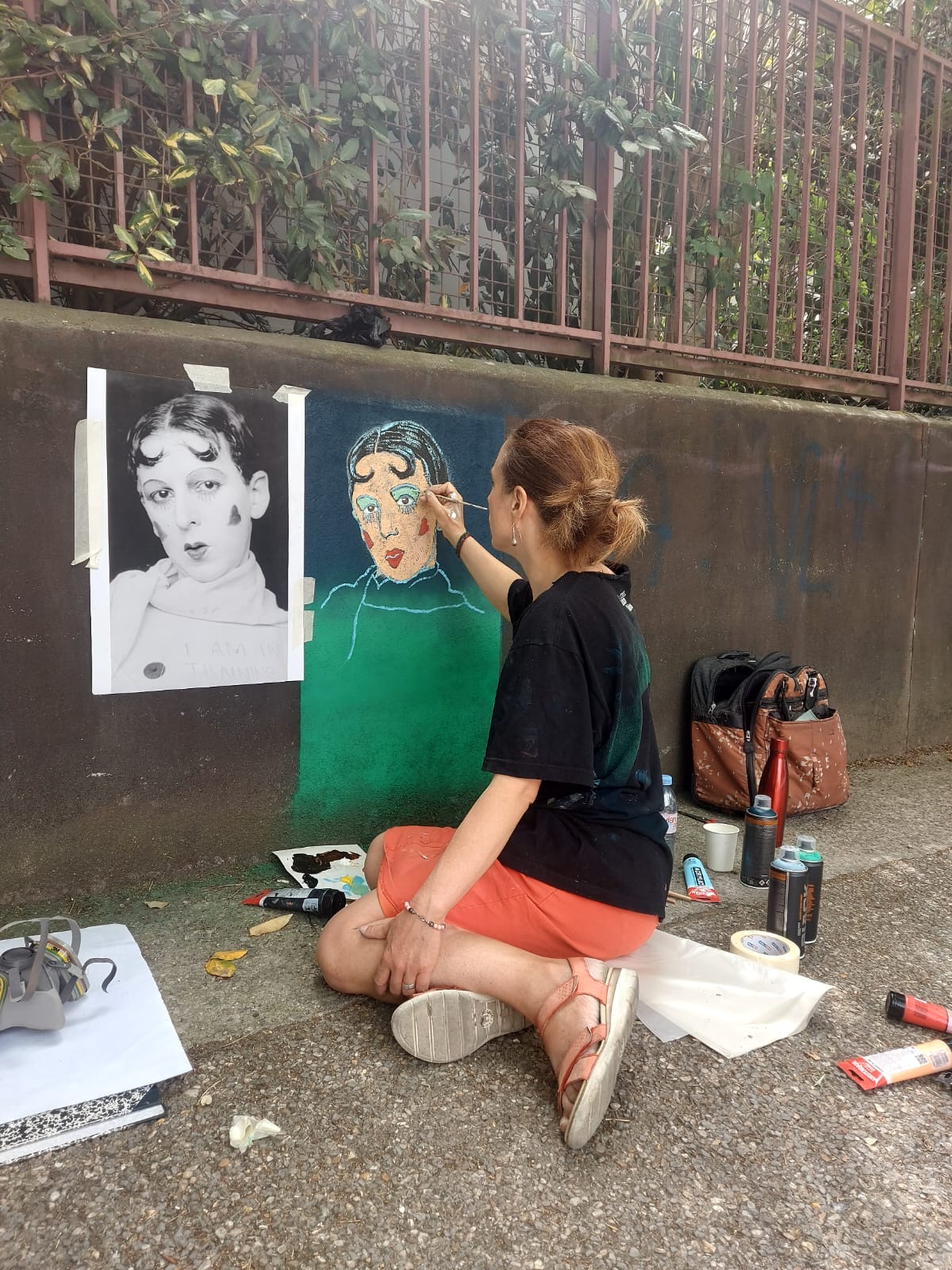 Clémence Devienne, assise sur le trottoir, peint au pinceau un portrait de Claude Cahun sur un mur, à côté d'une photographie de Claude Cahun