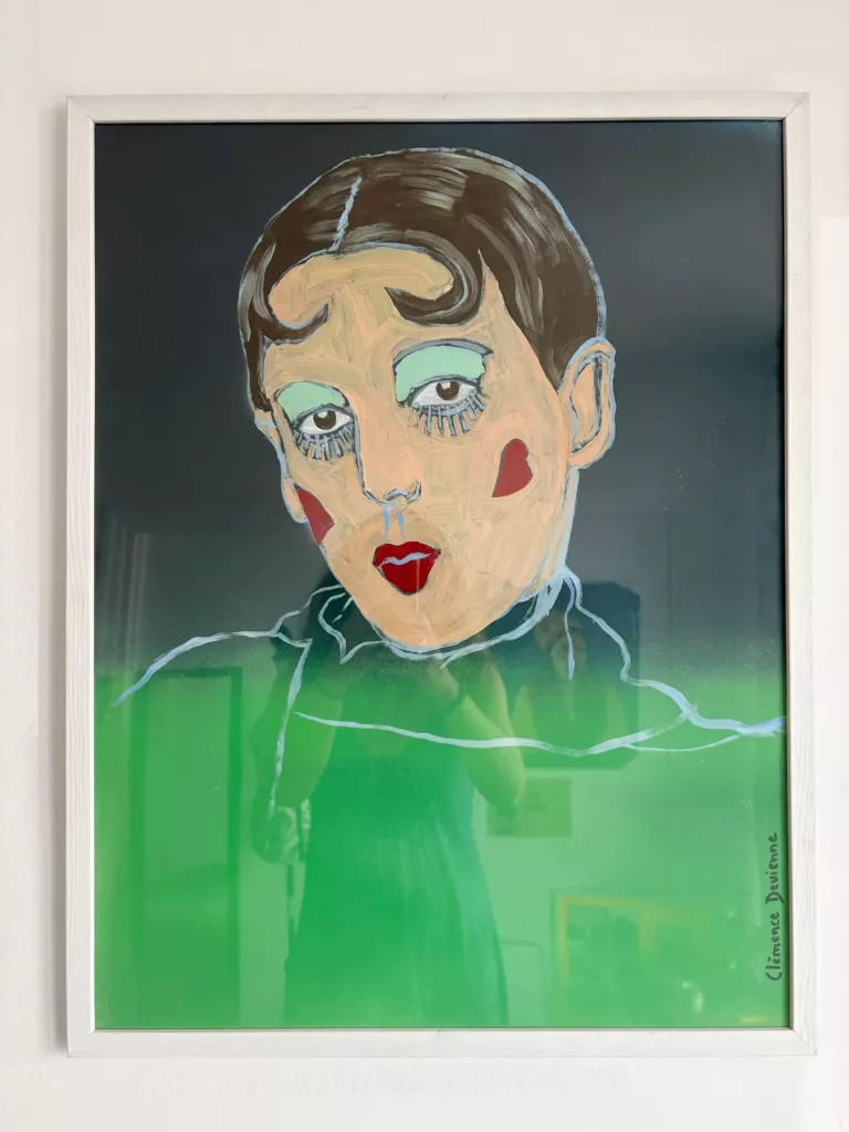 Portrait de Claude Cahun : visage pâle aux paupières vertes et aux joues rouges en forme de cœur, sur fond vert sombre, dans un cadre blanc