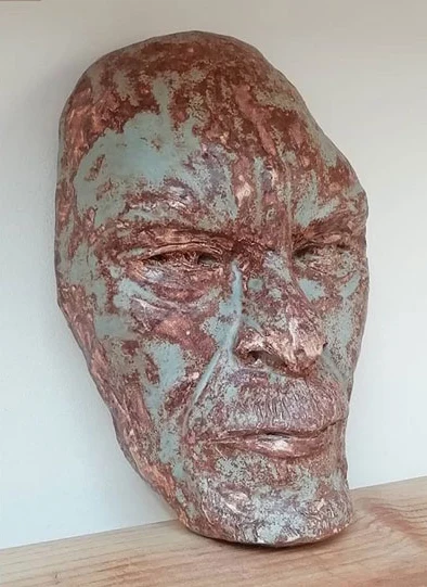 Tête d'Alfred Doucette en argile émaillée, vue de trois-quarts : visage marqué de rides, émail bleu-vert et cuivré, posée contre un mur blanc